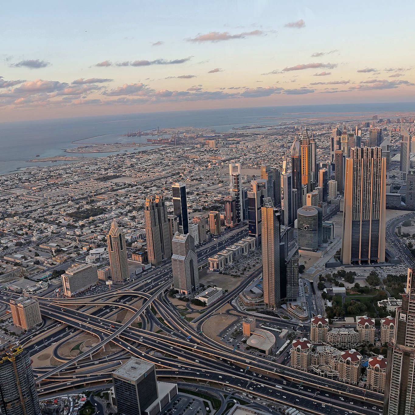 L'image montre un panorama impressionnant d'une ville moderne, probablement Dubai. On y voit de nombreux gratte-ciels élégants s'élevant vers le ciel, ainsi qu'un réseau complexe de routes et d'autoroutes serpentant à travers la métropole. À l'arrière-plan, on aperçoit la mer et des paysages urbains qui s'étendent à perte de vue. Le ciel présente des teintes douces de bleu et d'orange, témoignant d'un coucher de soleil, ce qui ajoute une ambiance tranquille à la scène urbaine animée.