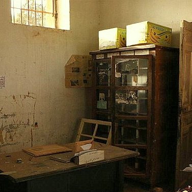 تظهر الصورة غرفة مهجورة مع جدران تحمل آثار الزمن، حيث تتواجد أريكة حمراء تقليدية على الجانبين. في الزاوية، يوجد مكتب خشبي بسيط بجانبه بعض الأوراق. على الجدار، توجد صورة معلقة لرجل يرتدي بدلة، مما يشير إلى أنه كان شخصية مهمة في الماضي. في الخلفية، يوجد زوج من خزائن الزجاج التي تحتوي على أشياء غير محددة. الإضاءة في الغرفة خافتة، مما يضيف إلى الأجواء الكئيبة للمكان.