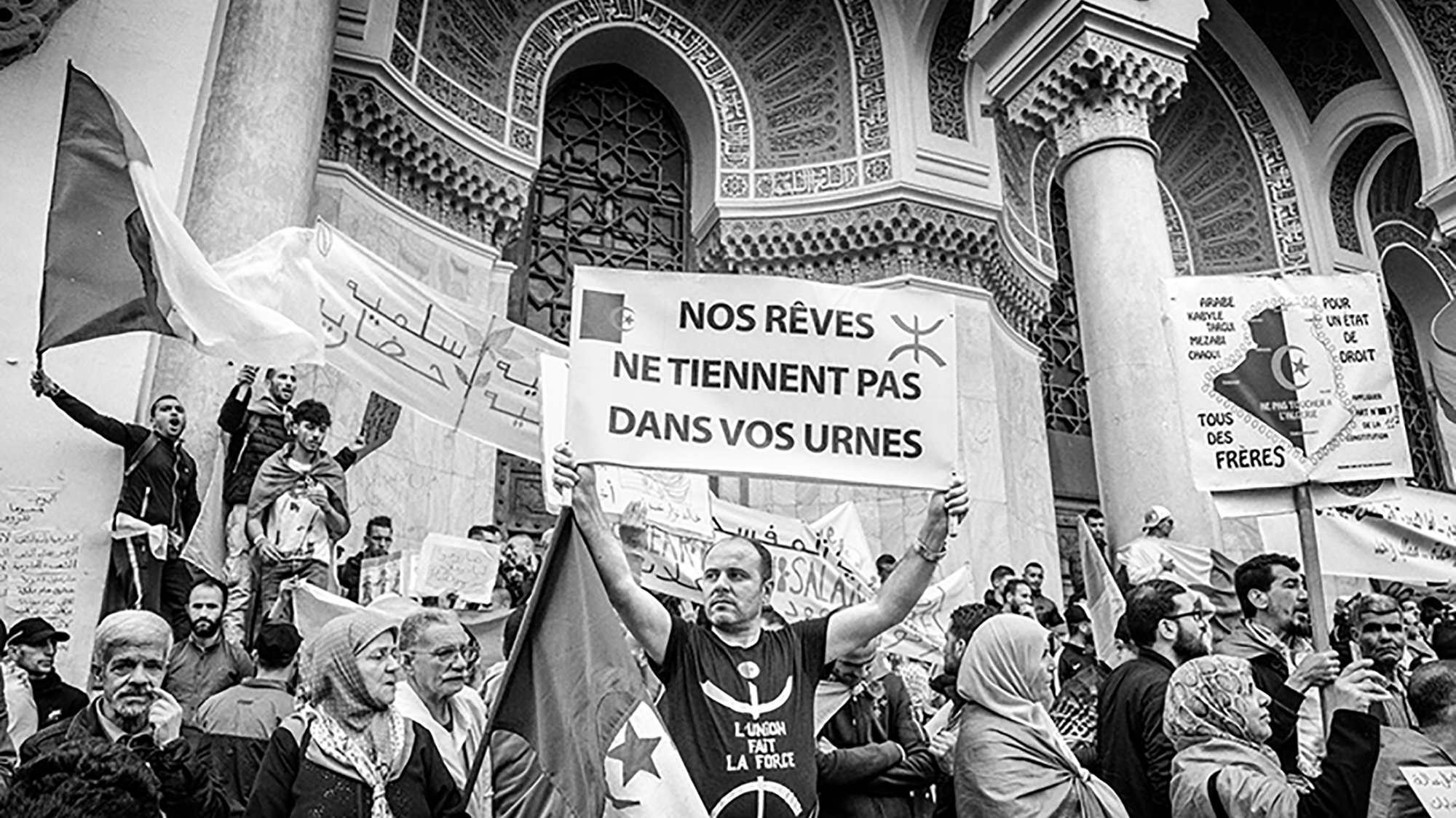 تظهر الصورة مجموعة من المتظاهرين في ساحة عامة، حيث يحمل أحدهم لافتة مكتوبًا عليها باللغة الفرنسية "أحلامنا لا تتواجد في صناديق الاقتراع". يُظهر المشهد حشدًا من الناس يتجمعون معًا، مع تباين واضح بين الأعمار والملابس. بعض الأشخاص يرتدون الأوشحة أو الحجاب، مما يعكس التنوع الثقافي. تعكس الأجواء إحساسًا قويًا بالتضامن والمشاركة في قضية معينة.