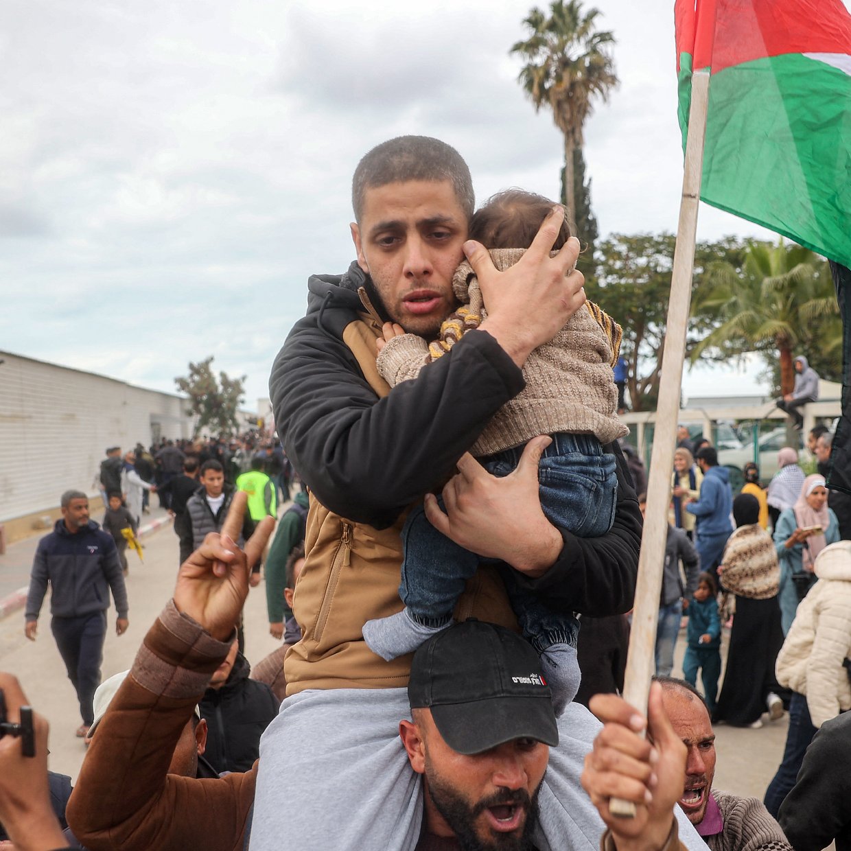 L'image montre une scène de rassemblement où un homme porte un enfant dans ses bras. Il semble vigilant et protecteur. En arrière-plan, il y a un groupe de personnes qui brandissent des drapeaux et expriment leur engagement ou leurs émotions. Des arbres et des structures sont visibles en arrière-plan, suggérant une ambiance de protestation ou de manifestation. L'atmosphère semble chargée en émotions.