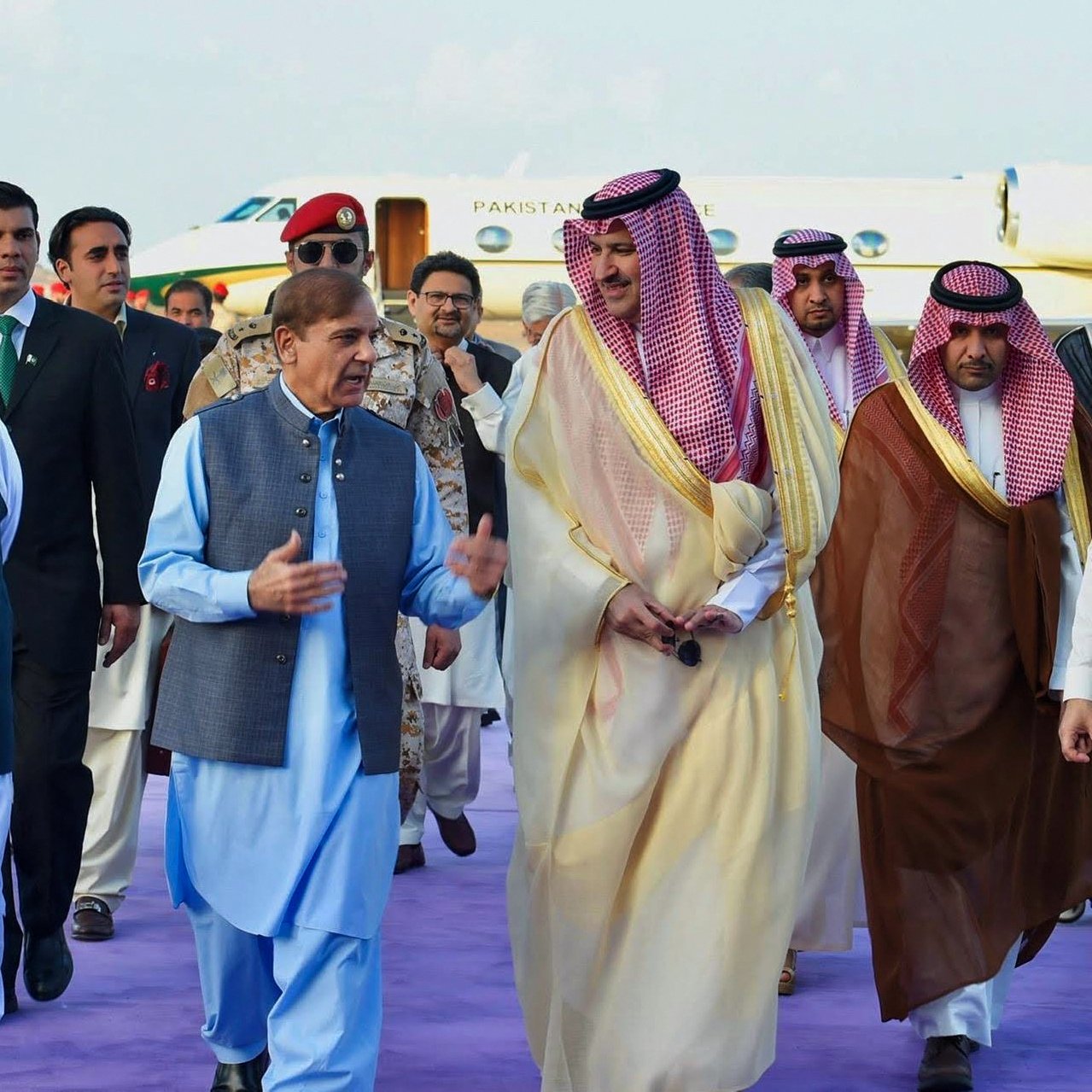 L'image montre un groupe de personnes en tenue formelle, marchant ensemble sur un tapis violet. On peut voir des hommes portant des costumes traditionnels et des vêtements d'apparat, avec certains en toges ou en robes. En arrière-plan, un avion privé est stationné, indiquant un contexte officiel ou diplomatique. L'atmosphère semble conviviale, avec les participants engagés dans une conversation.