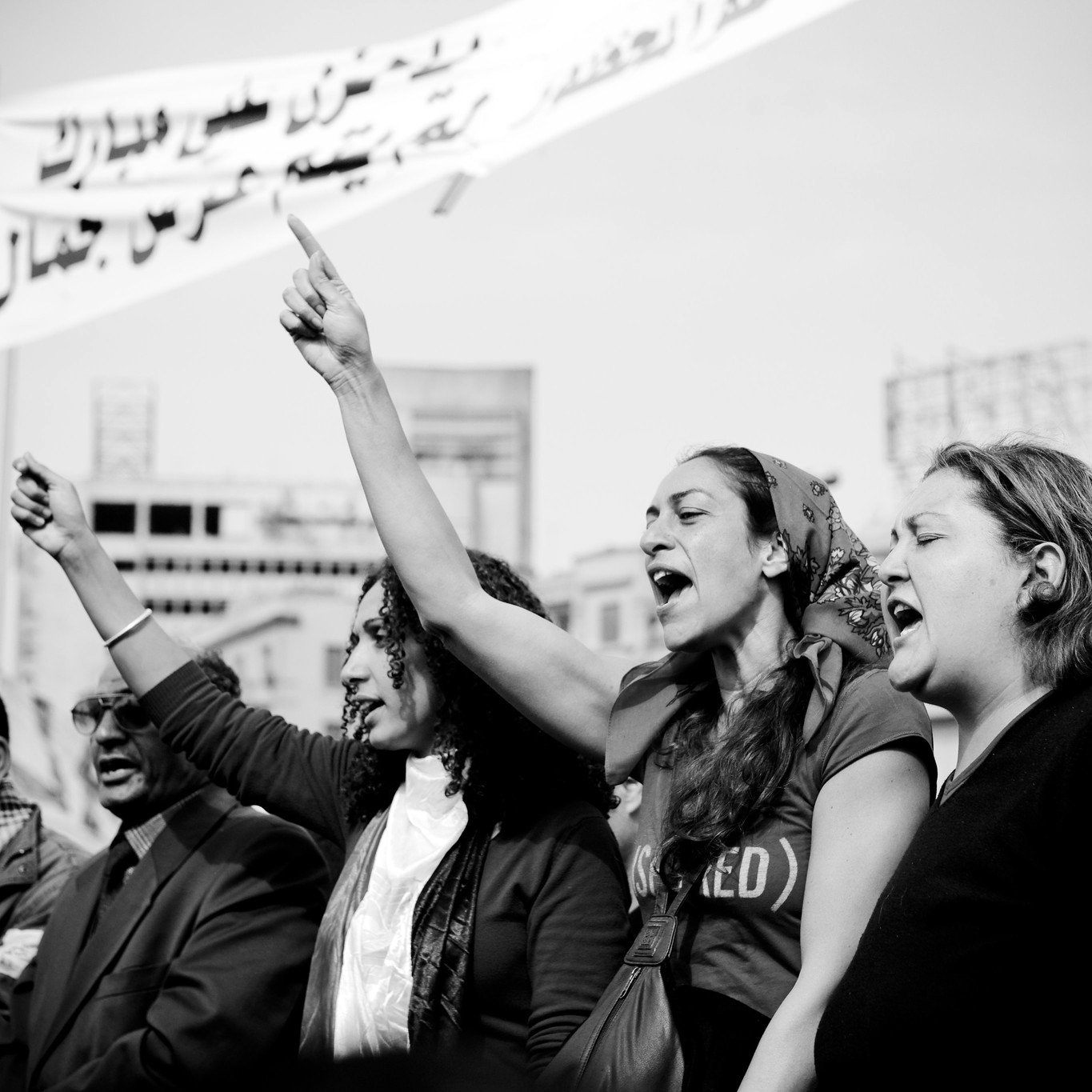 L'image montre un groupe de personnes participant à une manifestation. On peut voir des femmes au premier plan levant le poing et chantant, exprimant des émotions fortes et de la détermination. L'ambiance semble dynamique et engageante. En arrière-plan, des banderoles apparaissent, suggérant un message ou une demande collective. La photo est en noir et blanc, ce qui accentue l'intensité et l'impact visuel de la scène.