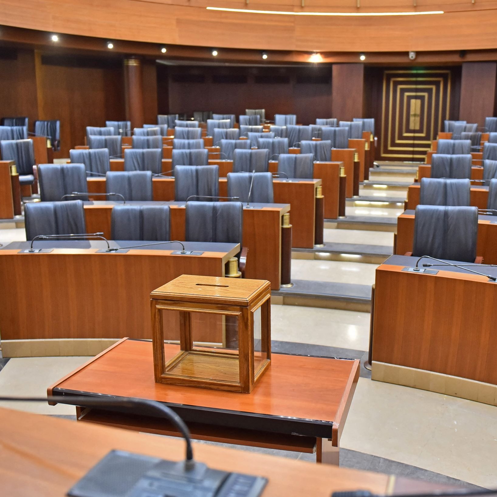 L'image montre une salle de réunion ou un parlement. Les sièges sont en bois, avec des dossiers noirs et sont disposés en rangées organisées. Au centre, il y a une petite table en bois avec une boîte en verre posée dessus. L'éclairage est doux, mettant en valeur les finitions en bois des murs et des meubles. L'ensemble de la salle dégage une atmosphère formelle et austère.
