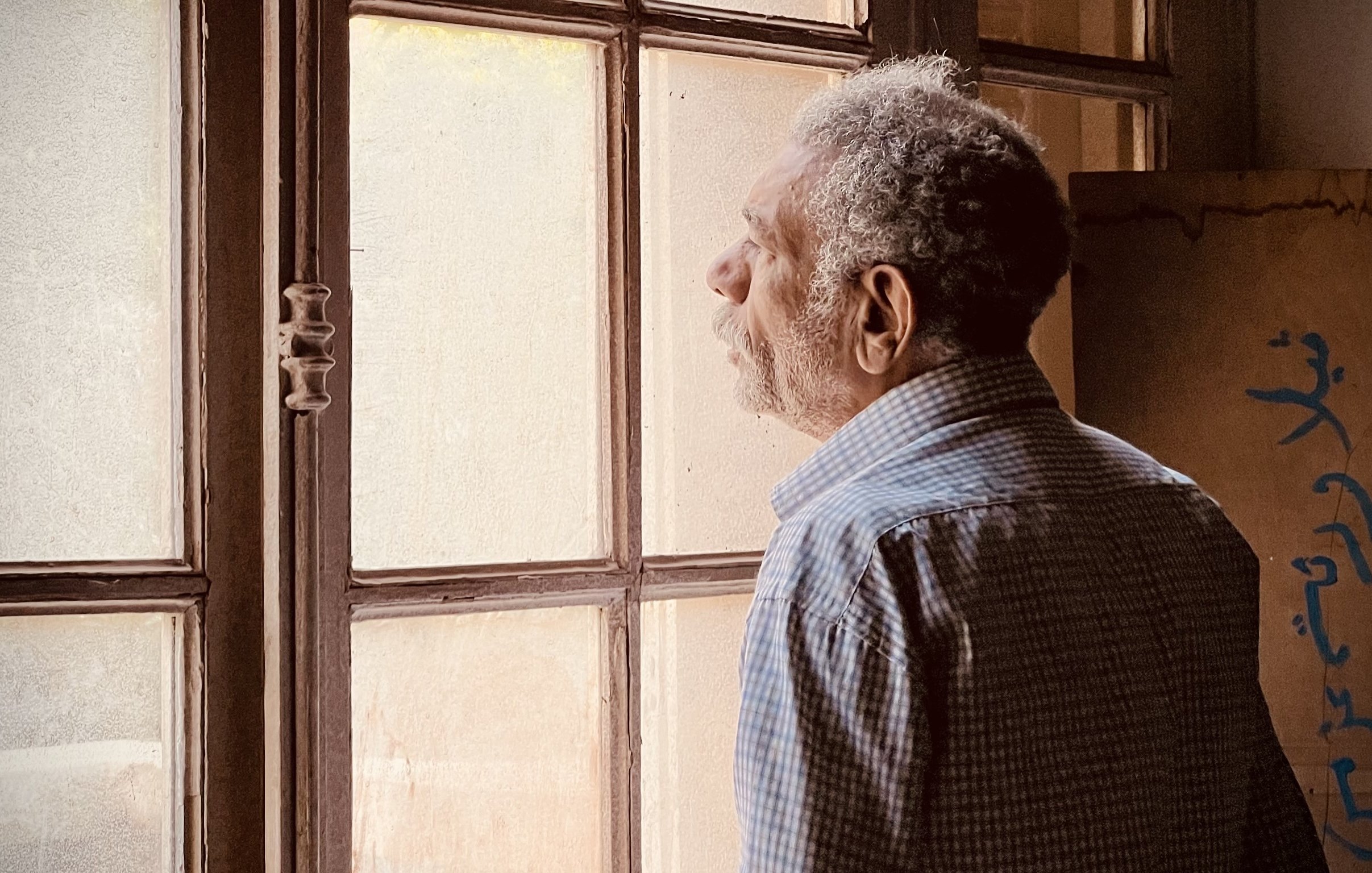 L'image montre un homme âgé qui semble contempler à travers une fenêtre. Il est habillé d'une chemise à motifs et a une posture pensive. La lumière naturalisée entre par la fenêtre, illuminant doucement son visage et créant une ambiance calme. On peut aussi apercevoir des murs en arrière-plan, ajoutant à l'atmosphère de réflexion du moment.