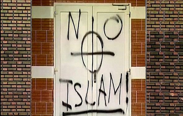 تظهر في الصورة بابًا مغطى بكتابات جدارية، حيث تم رسم عبارة "لا إسلام" مع رمز يظهر قوسًا دوائريًا. الكتابة تبدو باللون الأسود على خلفية بيضاء. الجدران المحيطة بالباب مصنوعة من الطوب بلون بني فاتح. الصورة تعكس مشاعر سلبية تجاه الإسلام، مما يثير جدلاً حول التسامح والقبول.