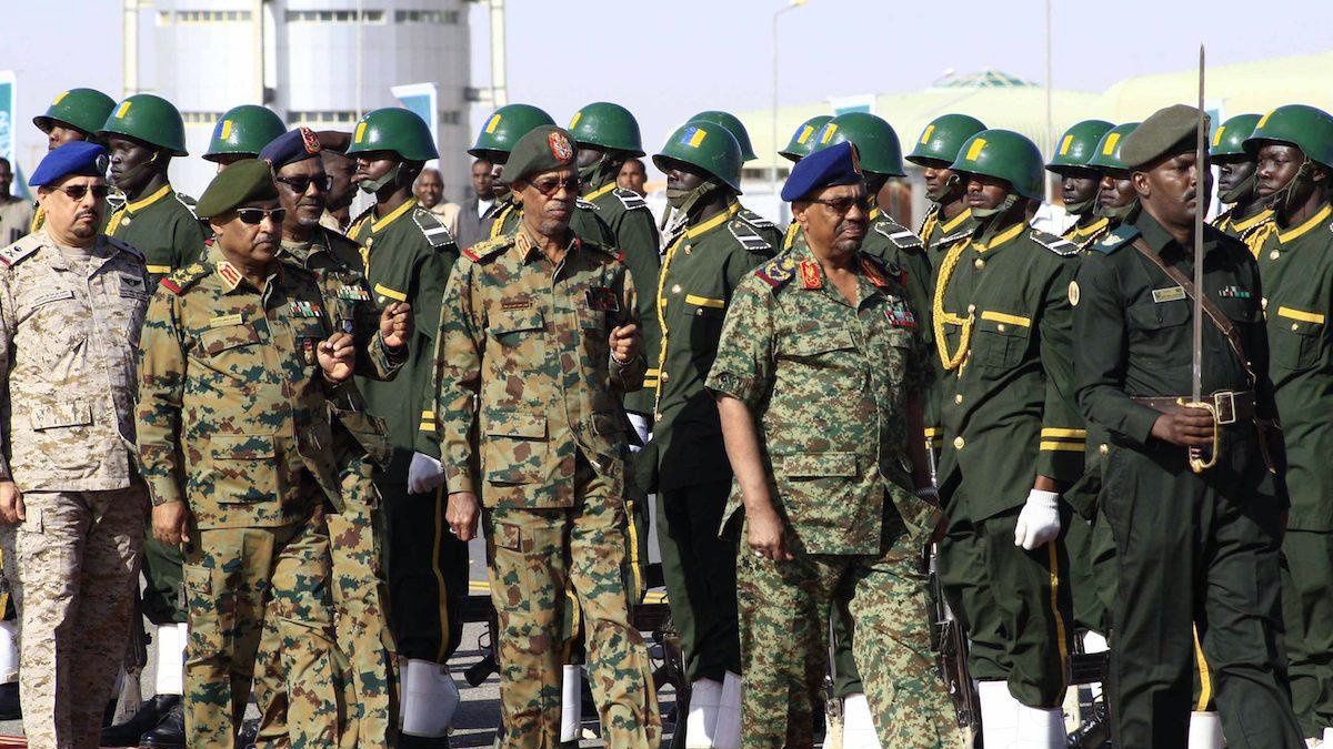 L'image montre un groupe de militaires en uniforme, marchant en formation. Les soldats portent des uniformes variés, notamment des uniformes camouflés et d'autres de couleur kaki, ornés de médailles et d'insignes. Certains militaires portent des casques ou des bérets. Au premier plan, des officiers semblent inspecter les troupes, tandis qu'une formation de soldats en arrière-plan est prête, alignée sur une surface rouge, probablement lors d'une cérémonie officielle. L'ambiance semble solennelle et rituelle, caractéristique d'une parade militaire.