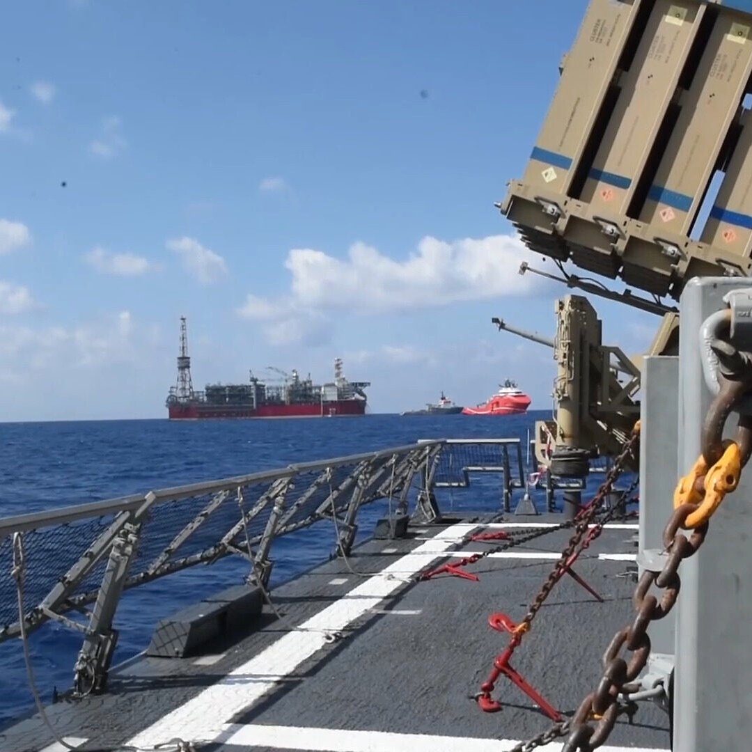 تظهر الصورة مشهدًا من سطح سفينة بحرية، حيث يمكن رؤية مجموعة من المنصات البحرية في البحر. المياه تبدو هادئة تحت سماء زرقاء مع بعض السحب البيضاء. من الجانب الأيسر، توجد معدات عسكرية أو دفاعية، ربما صواريخ أو أنظمة توجيه، بينما توجد منصات نفطية في الخلفية. تظهر أيضًا سفن أخرى على مسافة قريبة، مما يدل على نشاط بحري مكثف في المنطقة.