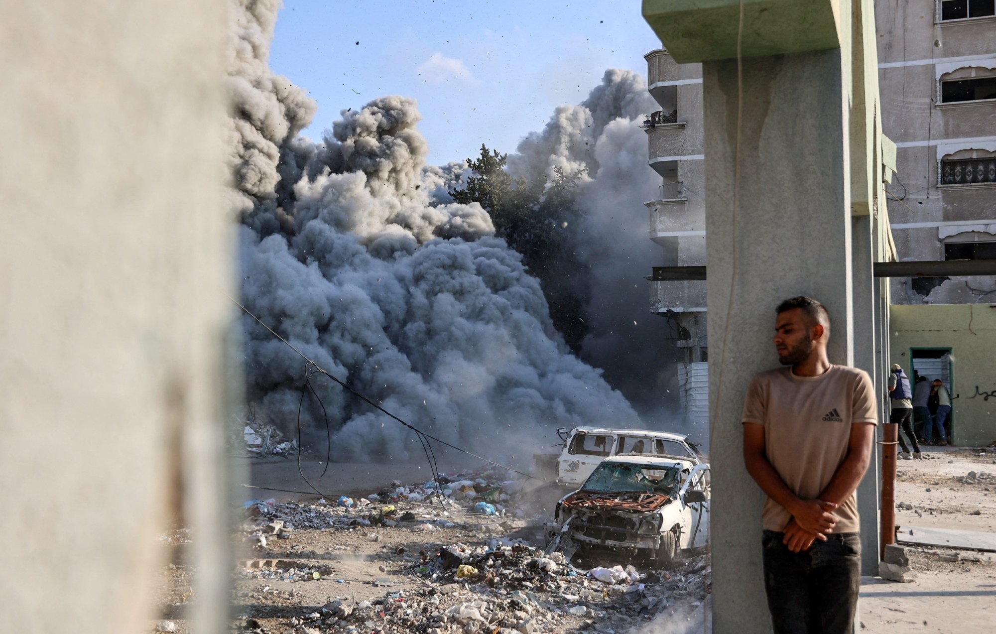 Destruction urbaine avec de la fumée, un homme adossé à un pilier près d'une voiture endommagée.