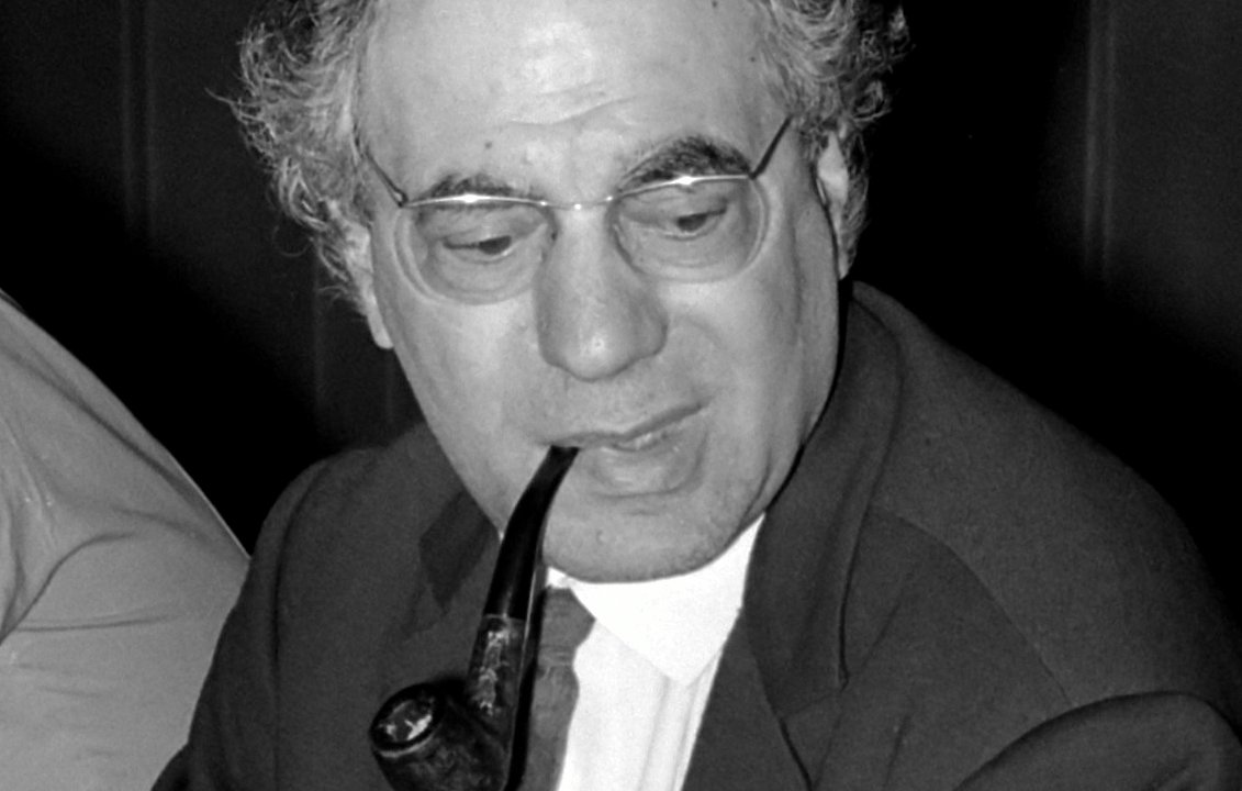 The image features a man seated at a table, wearing a suit and tie. He has a full head of curly hair and is holding a pipe in his mouth, which gives him a thoughtful or contemplative appearance. The setting seems to be a formal or professional environment, likely during a meeting or discussion. The lighting is subdued, which adds to the serious tone of the scene.