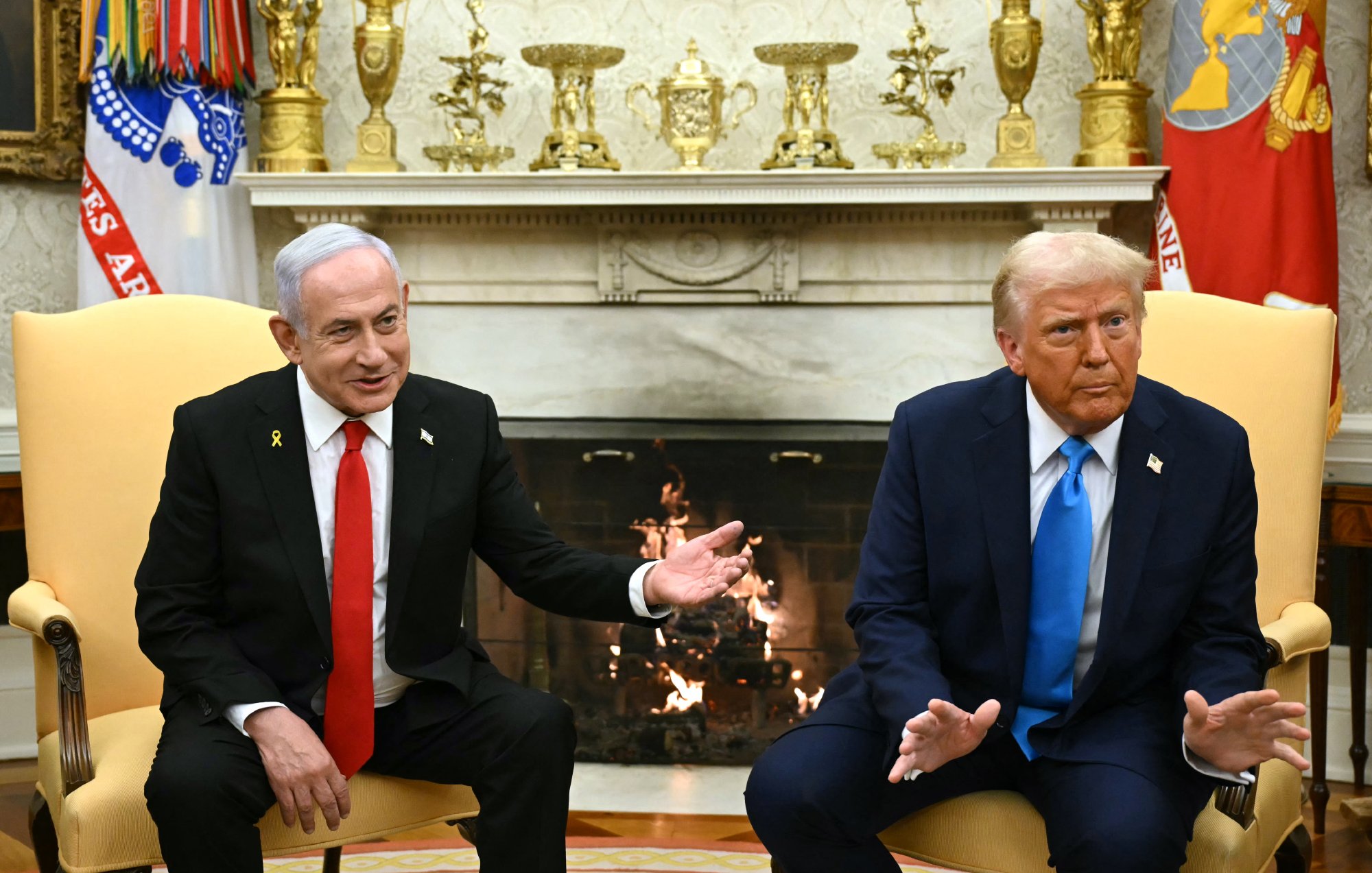 Nell'immagine si possono vedere due uomini seduti in poltrone di fronte a un camino. A sinistra c'è un uomo con una giacca nera, una cravatta rossa e un distintivo sul petto. A destra, l'altro uomo indossa un giubbotto blu e una cravatta rossa, anch'esso con un distintivo. Entrambi stanno parlando e sembrano impegnati in una conversazione. Sullo sfondo c'è un arredamento elegante, con decorazioni sulle pareti e un camino acceso.