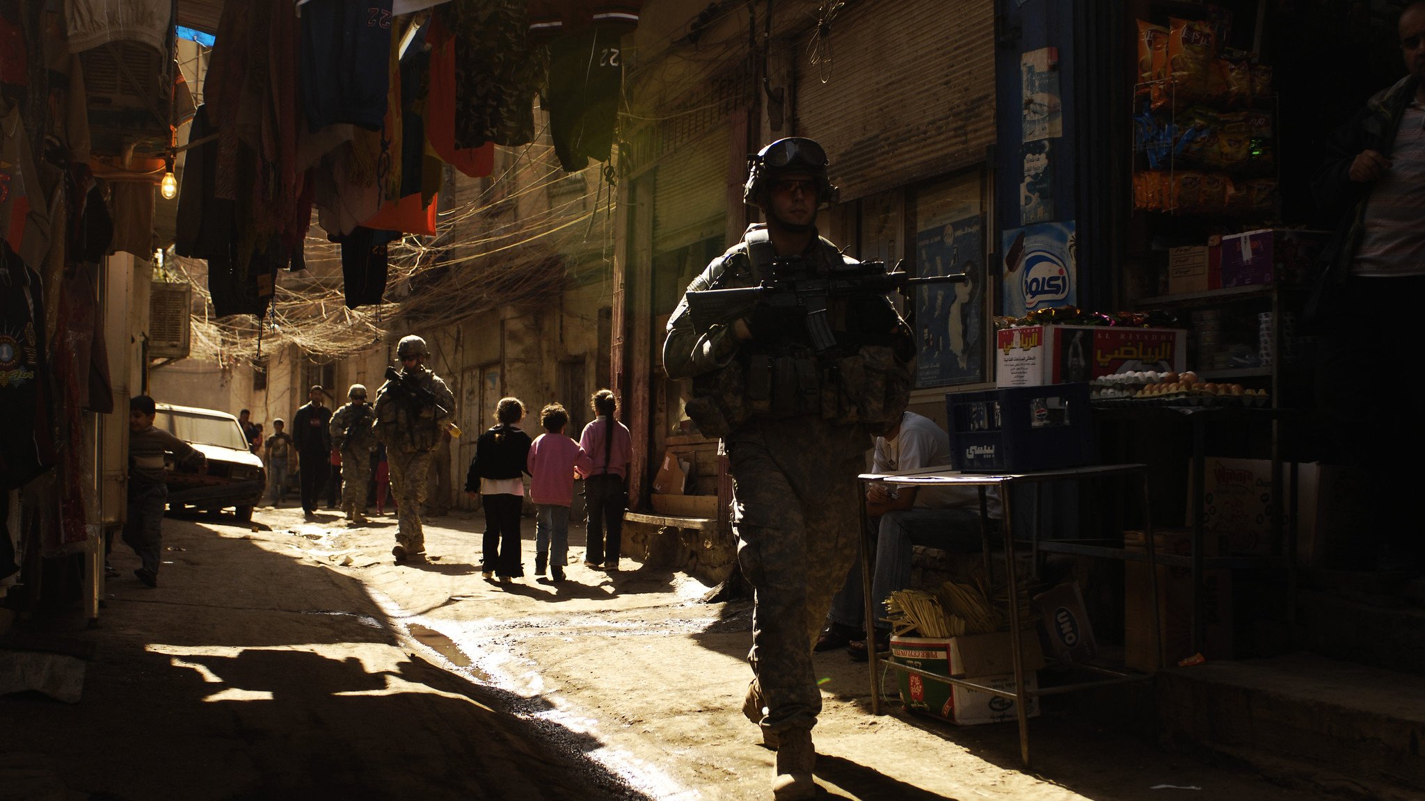 L'image montre une rue étroite et animée typique d'un marché urbain. Des soldats en uniforme, équipés d'armes, marchent le long de la ruelle. On aperçoit également des enfants qui semblent jouer ou observer les soldats. Les bâtiments sont proches les uns des autres, et des vêtements sont suspendus au-dessus de la rue. La lumière du soleil filtre à travers la ruelle, créant une ambiance contrastée entre l'ombre et la lumière. L'environnement semble à la fois vivant et chargé de tension.