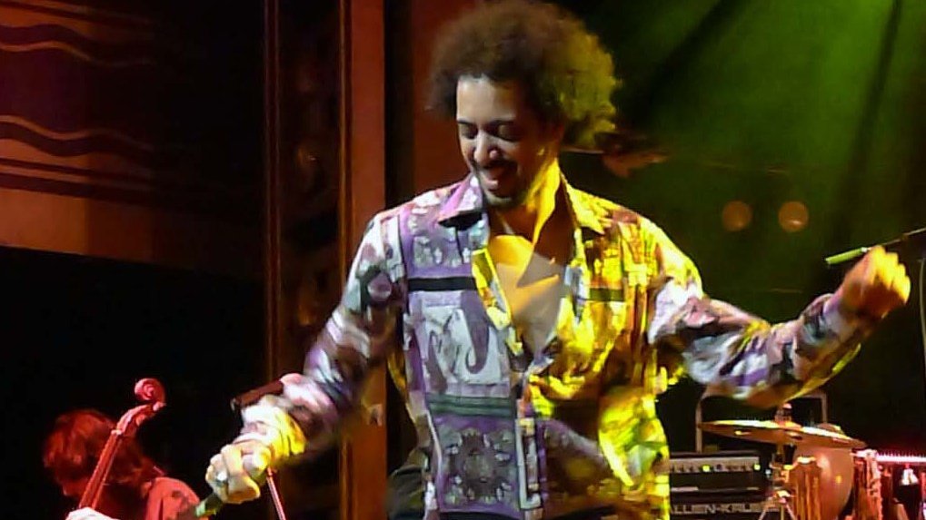 L'image montre un artiste en pleine performance sur scène. Il porte une chemise colorée à motifs et est en train de danser, manifestant une grande énergie. En arrière-plan, on peut apercevoir des musiciens jouant d'instruments, ce qui suggère un concert en direct. L'éclairage de la scène crée une atmosphère dynamique et festive.