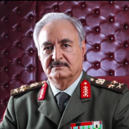 L'image montre un homme en tenue militaire, assis sur une chaise dans un cadre élégant. Il porte un uniforme avec des épaulettes ornées de médailles et d'insignes. L'arrière-plan est composé d'un panneau rembourré en cuir de couleur rouge, ce qui ajoute une touche de sophistication à l'ensemble. L'expression de l'homme est sérieuse et confiante.