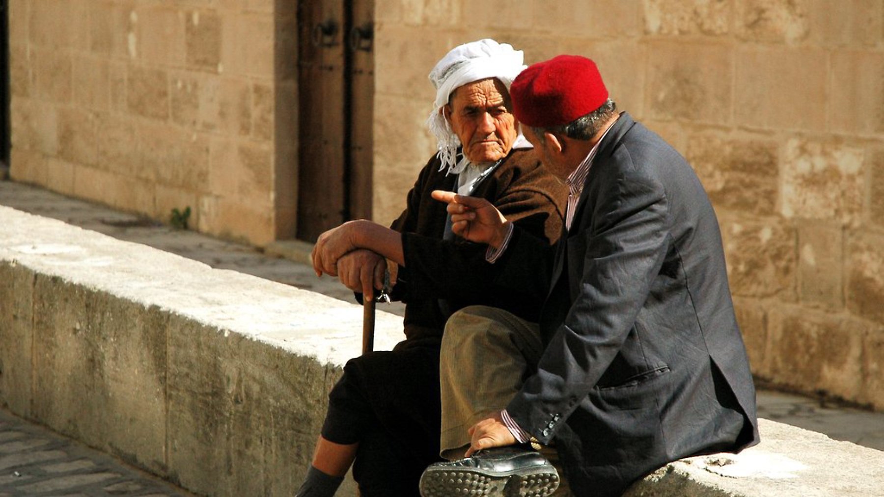 Deux hommes âgés discutent assis sur un mur en pierre, l'un portant un fez rouge.