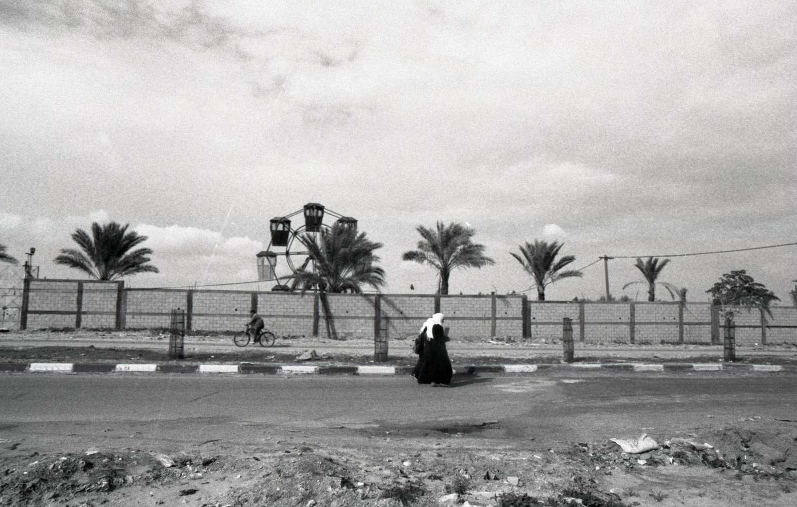 L'immagine ritrae una scena in bianco e nero che mostra una figura solitaria che cammina lungo una strada. Sullo sfondo, si possono vedere alcune palme e una ruota panoramica, che suggerisce la presenza di un parco o di un'area ricreativa. La composizione offre un'atmosfera malinconica e riflessiva, accentuata dalla scelta dei toni scuri e dalla mancanza di colori vivaci. La figura, vestita con abiti tradizionali, sembra essere in un momento di solitudine all'interno di un paesaggio urbano.