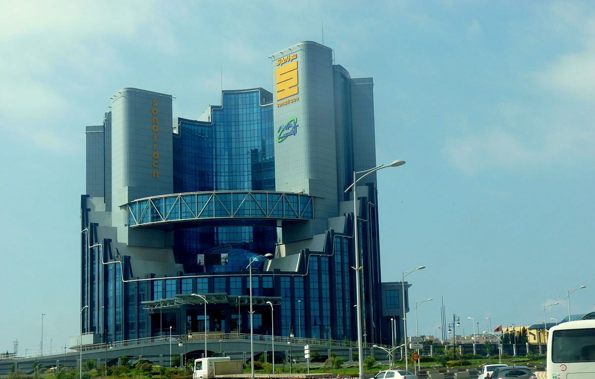 L'image montre un bâtiment moderne et imposant, avec une architecture contemporaine. La structure se dresse sur plusieurs niveaux, avec de grandes fenêtres en verre qui reflètent la lumière. Au sommet, il y a un panneau avec des inscriptions en caractères distincts, suggérant que le bâtiment abrite une entreprise ou une organisation importante. Il est entouré d'une route et d'un espace vert, ce qui ajoute au cadre urbain. Le ciel est partiellement nuageux, ce qui donne un éclairage intéressant à l'ensemble de la scène.
