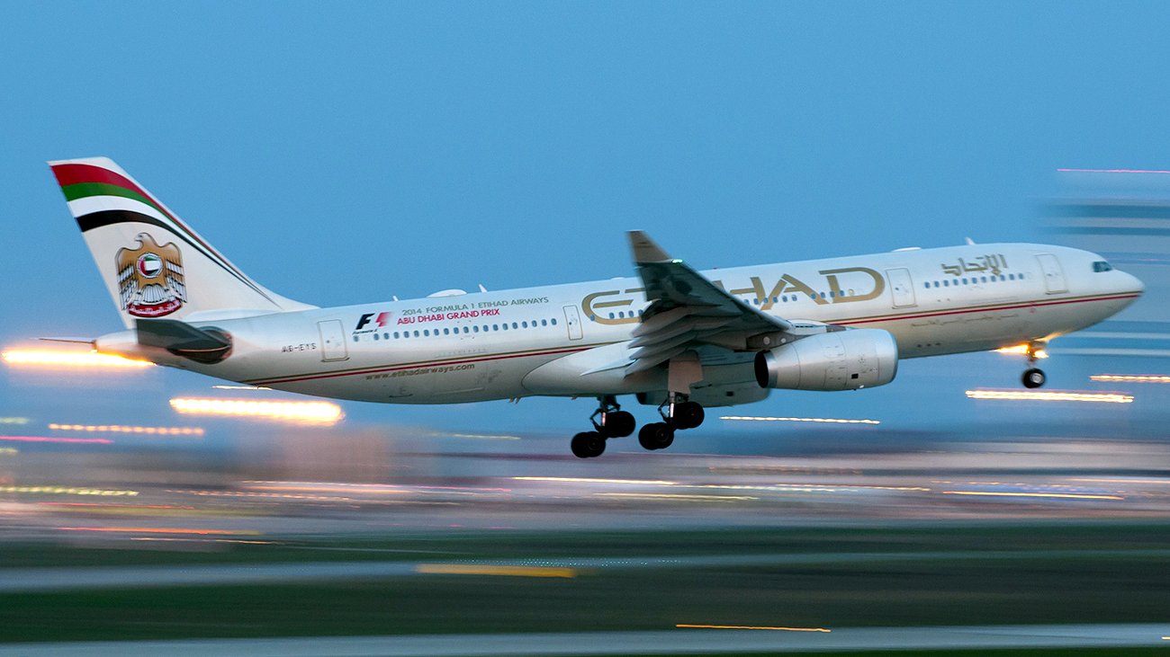 L'image montre un avion de la compagnie aérienne Etihad Airways en phase de décollage ou d'atterrissage. L'avion est partiellement flou, ce qui suggère une vitesse élevée. Le ciel apparaît dans une teinte bleue, tandis que des lumières brillantes en arrière-plan ajoutent une ambiance dynamique. L'appareil affiche la décoration et le logo caractéristiques de la compagnie.