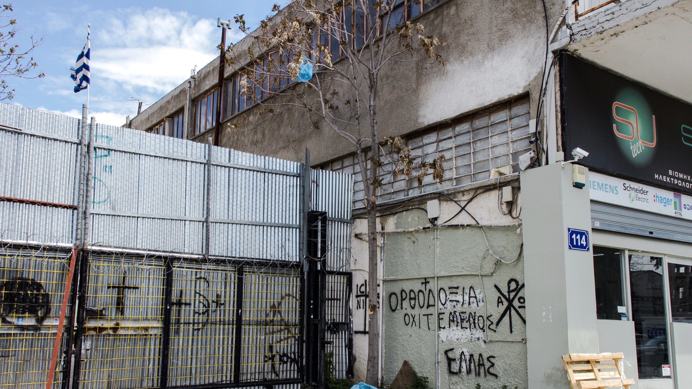 L'image montre un environnement urbain avec un bâtiment en arrière-plan et un mur métallique en façade, où l'on peut voir des graffitis. Un drapeau grec est visible, ainsi qu'un espace de stationnement ou de stockage à côté d'une entrée. Le sol semble en béton, et quelques déchets sont éparpillés sur le sol. Les graffitis sur le mur comprennent des inscriptions en grec, ce qui suggère un contexte local ou culturel. L'atmosphère paraît un peu désordonnée, typique des zones industrielles ou abandonnées.