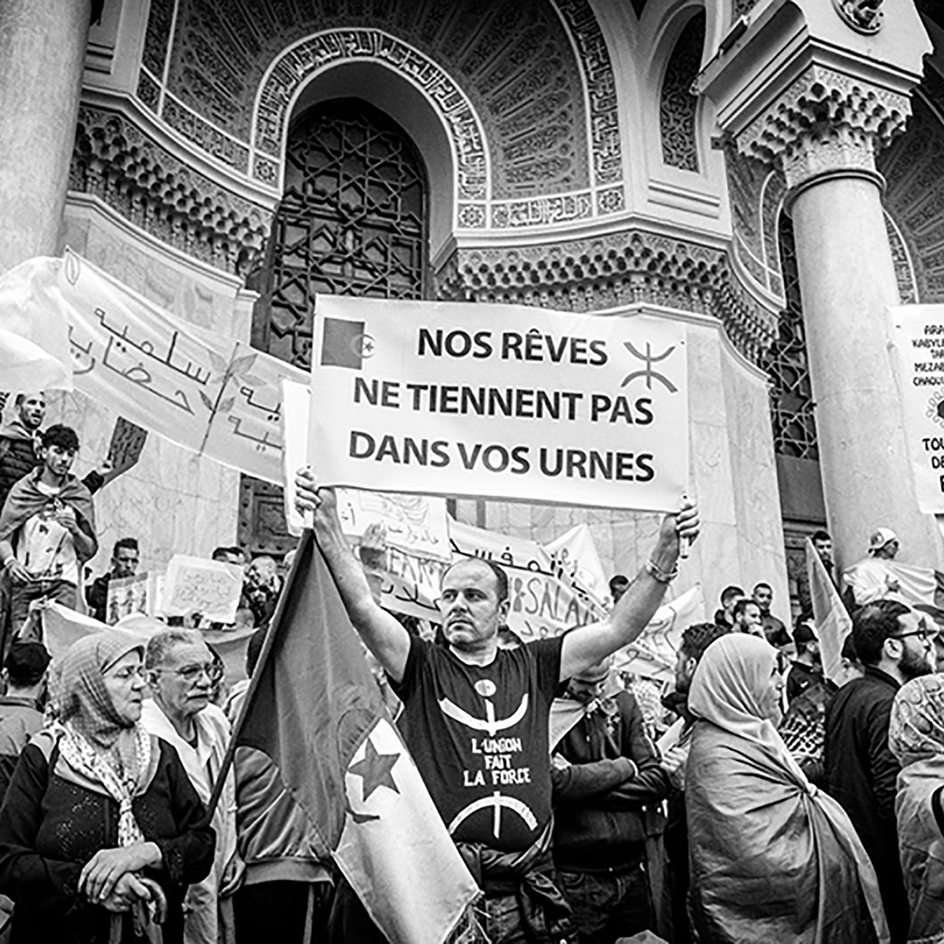 L'image montre une scène de manifestation où plusieurs personnes sont rassemblées. Au premier plan, un homme tient une grande pancarte qui affiche le message : "NOS RÊVES NE Tiennent PAS DANS VOS URNES". Les manifestants entourant cet homme expriment des sentiments de protestation, avec des drapeaux et des banderoles visibles. L'atmosphère semble chargée d'émotion et d'engagement politique, le tout dans un cadre architectural imposant. La photo est en noir et blanc, ce qui accentue l'intensité de la situation.