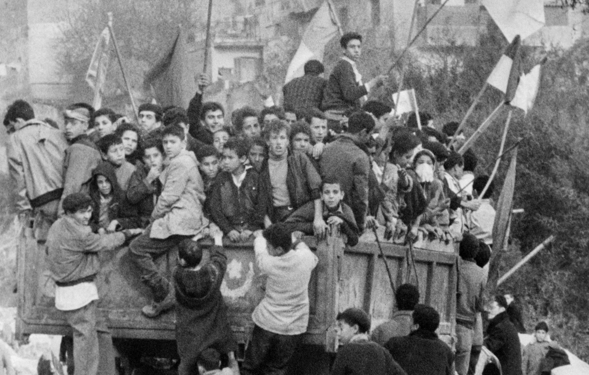 L'image montre un groupe de jeunes garçons et d'adolescents rassemblés sur un camion, entourés de drapeaux. Ils semblent célébrer un événement, peut-être une manifestation ou une fête. Les enfants sont penchés et accrochés au camion, reflétant un moment de camaraderie ou d'excitation collective. En arrière-plan, on aperçoit des bâtiments typiques d'une zone urbaine, contribuant à l'ambiance dynamique de la scène.