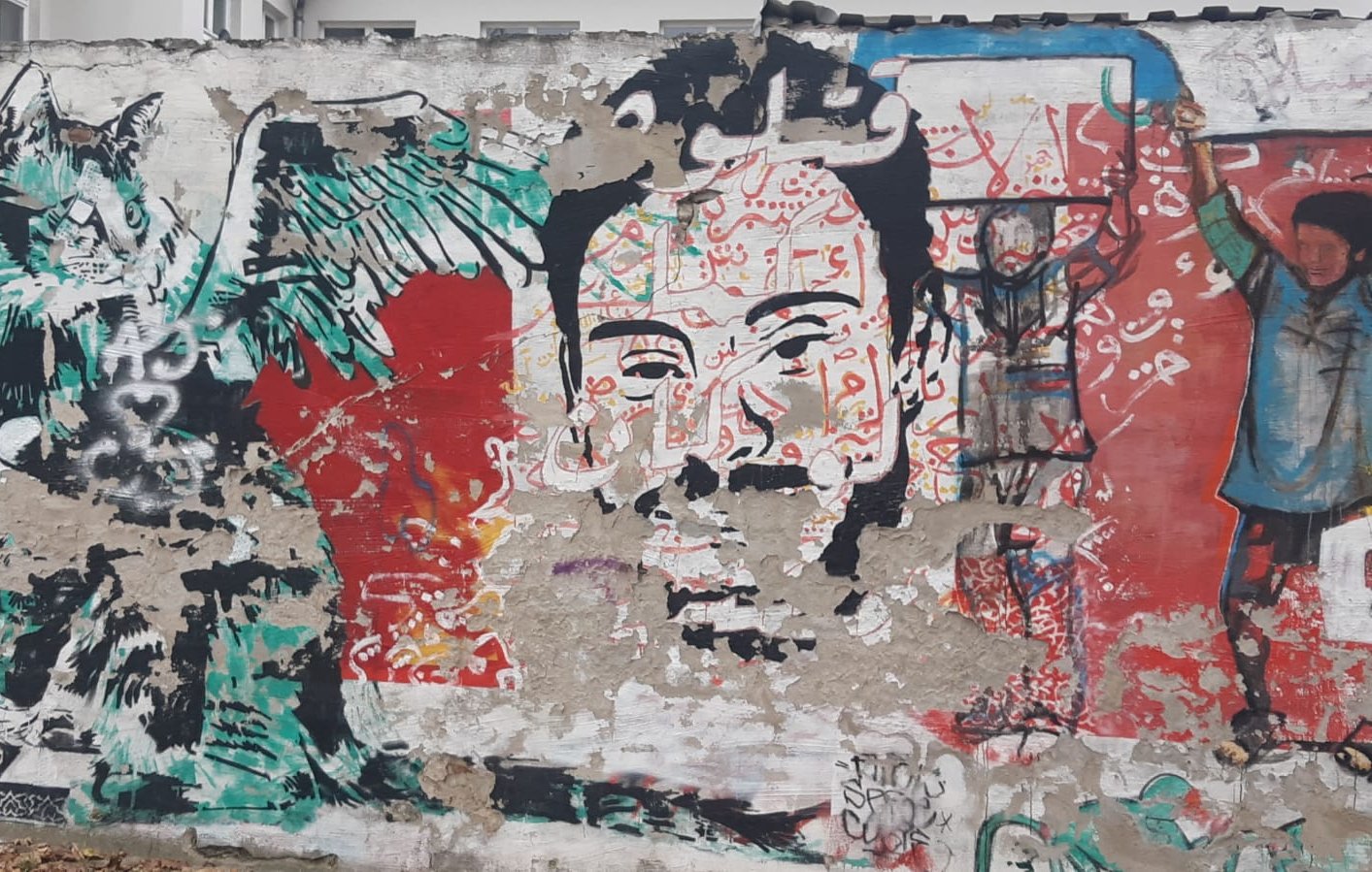 L'image montre une fresque murale riche en couleurs et en détails. À gauche, on peut voir un personnage avec des ailes et un visage expressif, qui semble être une représentation stylisée d'une créature. À droite, se trouve un visage humain qui attire l'attention, entouré de motifs vibrants. En arrière-plan, divers éléments graphiques et des inscriptions en arabe ajoutent une dimension culturelle. L'ensemble donne une impression de dynamisme et de créativité, typique de l'art urbain.