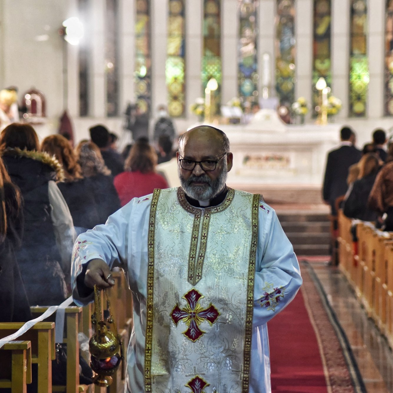 A priest in white robes walks down the aisle of a church during a service.