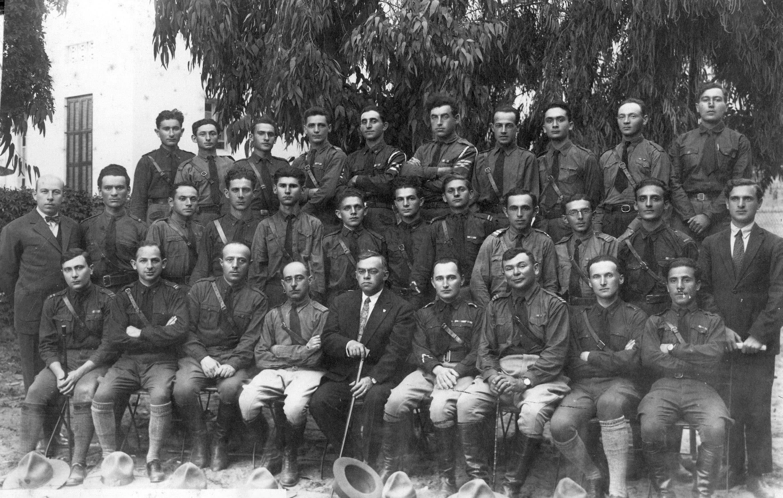 L'image montre un groupe de soldats en uniforme, probablement pris au début du 20e siècle. Ils sont assis et debout en rangée, avec des expressions sérieuses. Au centre, un homme en costume, tenant une canne, semble superviser le groupe. L'arrière-plan présente des arbres feuillus, ajoutant une touche naturelle à la scène. L'ambiance générale évoque une certaine solennité et un sentiment de camaraderie parmi les membres du groupe.
