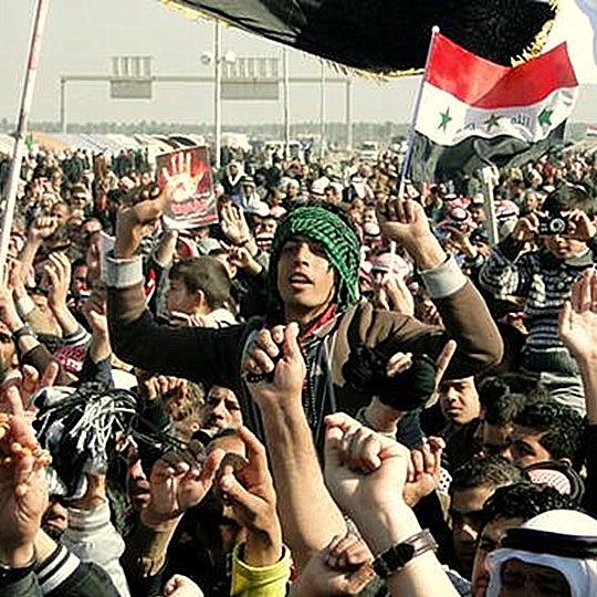 L'image montre une grande foule rassemblée, probablement lors d'une manifestation ou d'un événement public. Les personnes levées les bras, brandissant des drapeaux, affichent des expressions d'énergie et d'engagement. Certains portent des vêtements traditionnels, et l'ambiance semble dynamique et fervente. Des pancartes ou des symboles visibles pourraient suggérer un message politique ou social. L'environnement semble être un espace ouvert, peut-être une place ou une route.