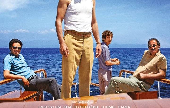 L'image montre quatre hommes sur un bateau, prenant la pose avec un fond marin. Le personnage central, portant un débardeur blanc, semble confiant et déterminé, tandis que les autres hommes sont assis autour de lui. Leurs expressions reflètent une atmosphère d'espoir et de camaraderie. Le texte mentionne "Algérie 1962, l'avenir est devant eux" et indique qu'il s'agit d'un film intitulé "L'Oranais", réalisé par Lyes Salem.