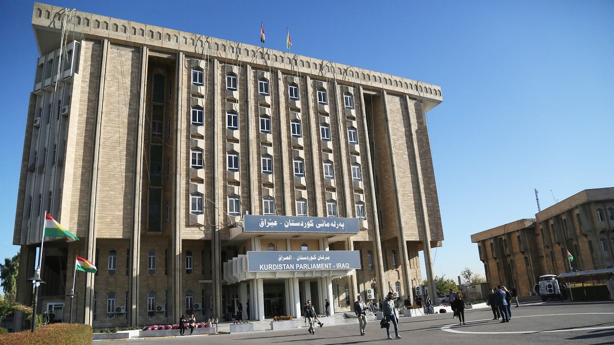 L'image montre un bâtiment imposant, probablement un édifice institutionnel, avec des drapeaux flottant à l'extérieur. La façade du bâtiment est en pierre avec un design moderne, et il est clairement identifié par une inscription en arabe et en anglais indiquant qu'il s'agit du "Parlement du Kurdistan - Irak". Il y a des personnes qui se déplacent autour du bâtiment, ce qui évoque un lieu d'activité politique et administrative. L'environnement semble clair et ensoleillé, avec un ciel bleu en arrière-plan.