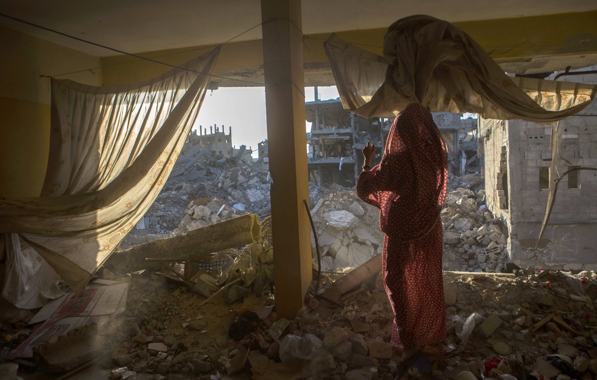 L'image montre une scène à l'intérieur d'un bâtiment endommagé. On aperçoit une femme vêtue d'un vêtement traditionnel, se tenant près d'une fenêtre qui donne sur des ruines. De l'autre côté, on voit des décombres de bâtiments effondrés, laissant entrevoir la désolation d'un environnement détruit. La lumière du soleil filtre à travers les rideaux, ajoutant une ambiance à la fois triste et poignante à la scène.