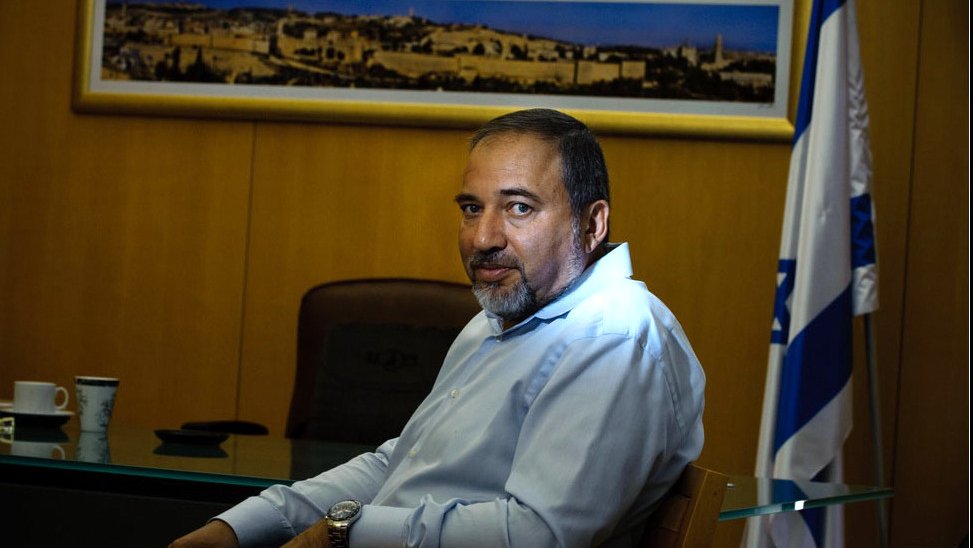 L'image montre un homme assis à un bureau. Il porte une chemise claire et a une expression sérieuse. En arrière-plan, on peut voir une grande photo d'une ville, probablement une vue de Jérusalem, et un drapeau, suggérant un contexte officiel ou politique. Le décor est sobre, avec une table en verre et une tasse sur le bureau.