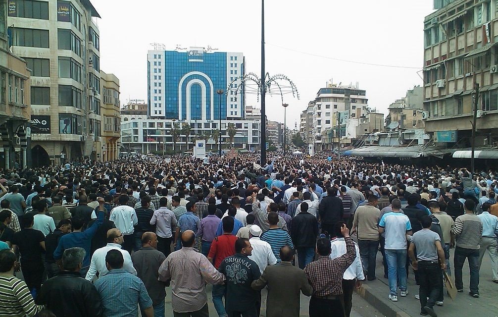 L'image montre une grande foule de personnes rassemblées dans une zone urbaine. On peut voir des bâtiments modernes en arrière-plan, ainsi qu'un aménagement urbain typique avec des lampadaires. La multitude semble engagée dans une manifestation ou un rassemblement, avec des gens de différentes orientations et tenues. L'ambiance générale évoque un moment de solidarité ou de protestation.