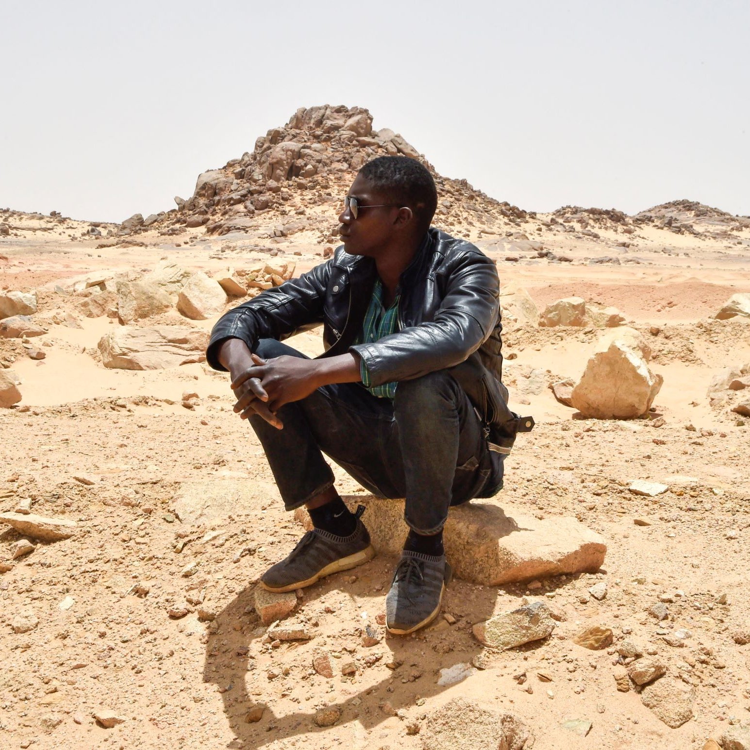 تظهر الصورة رجلين يجلسان في منطقة صحراوية. أحدهما يجلس على صخرة ويبدو عليه التفكير، بينما الآخر يجلس في الخلف بعيدًا قليلاً. الخلفية تتكون من صخور وكثبان رملية تحت سماء صافية. يبدو المشهد هادئًا، مما يعكس طبيعة الصحراء القاحلة.