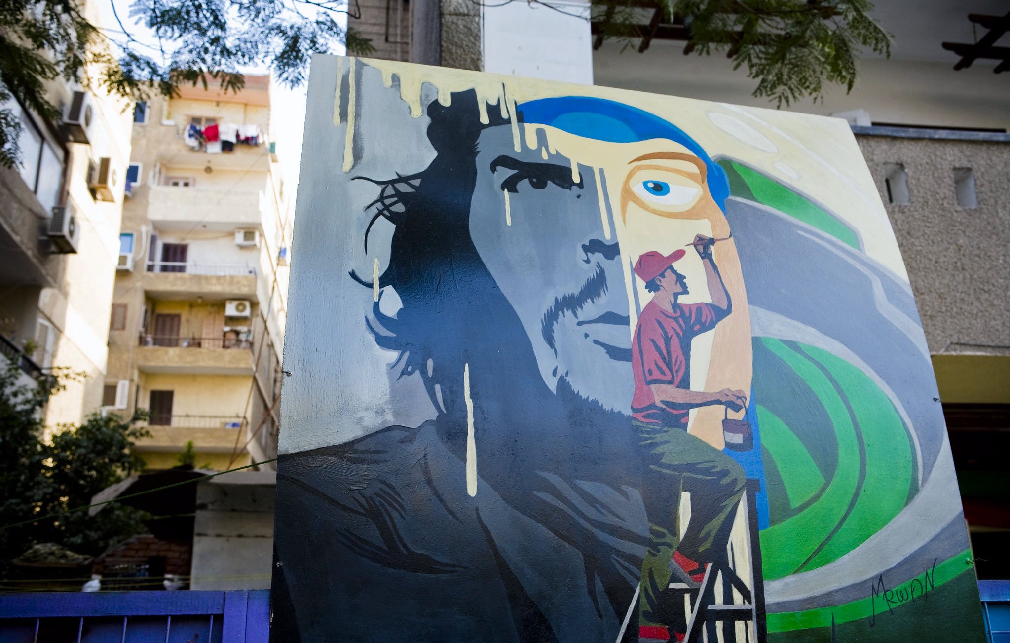 L'immagine mostra un murale artistico su un muro esterno. Il dipinto raffigura il volto di un uomo con un'espressione intensa, accanto a un artista in azione, intento a dipingere. Le tonalità utilizzate sono vivaci, con predominanza di blu e verde, creando un contrasto interessante. Sullo sfondo, si intravedono elementi architettonici e tende, suggerendo un ambiente urbano. Questo murale esprime una fusione tra arte e quotidianità.