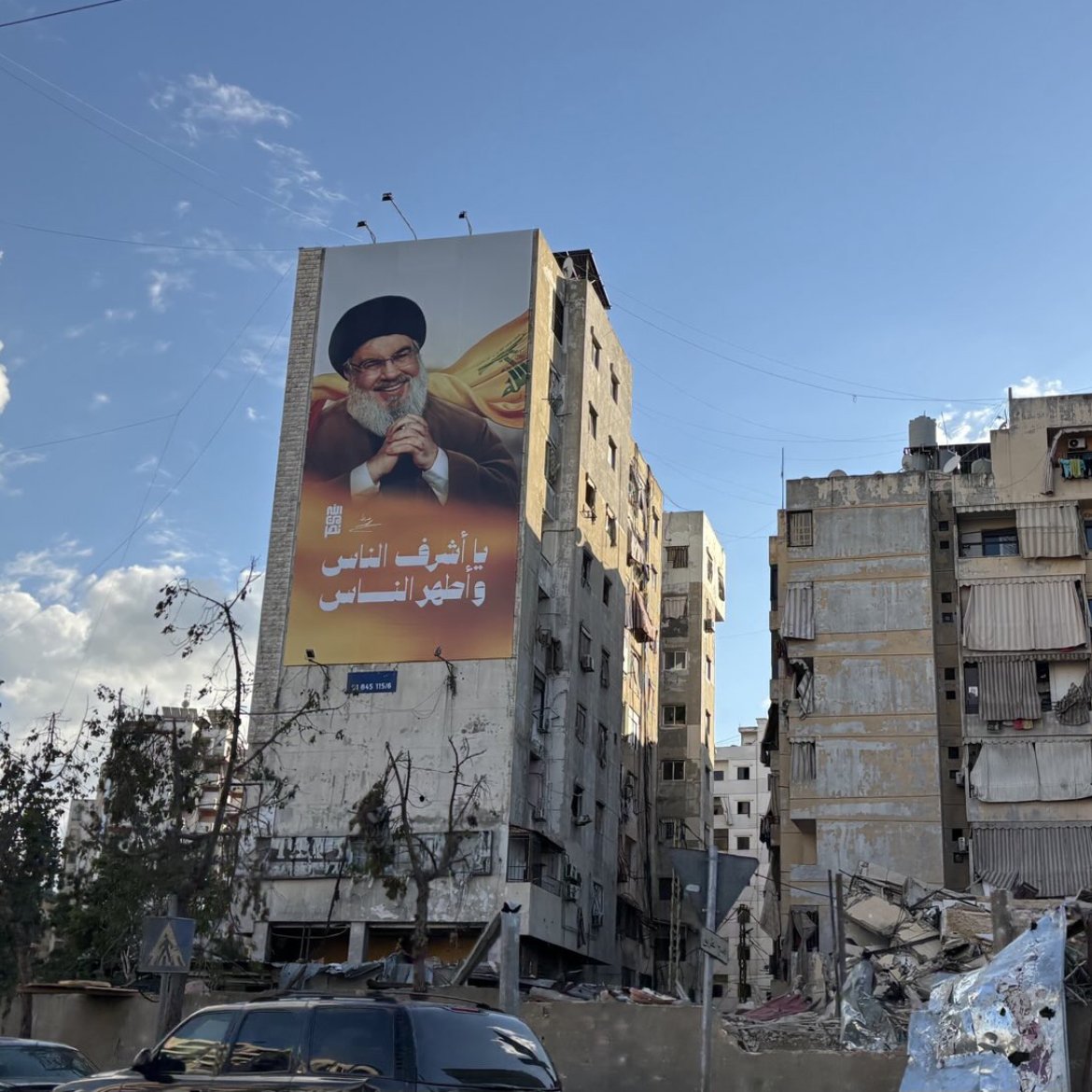 L'image montre un bâtiment en ruines, probablement à la suite d'un conflit ou d'une catastrophe, avec un autre bâtiment voisin encore debout mais en mauvais état. Sur le mur du bâtiment en meilleur état, il y a une grande affiche représentant un homme, probablement un leader ou une figure politique, tenant un symbole de pouvoir. Le ciel est partiellement nuageux, ce qui donne une ambiance contrastée à la scène. Les structures montrent des signes de délabrement, avec des fenêtres manquantes et des balcons en désordre.