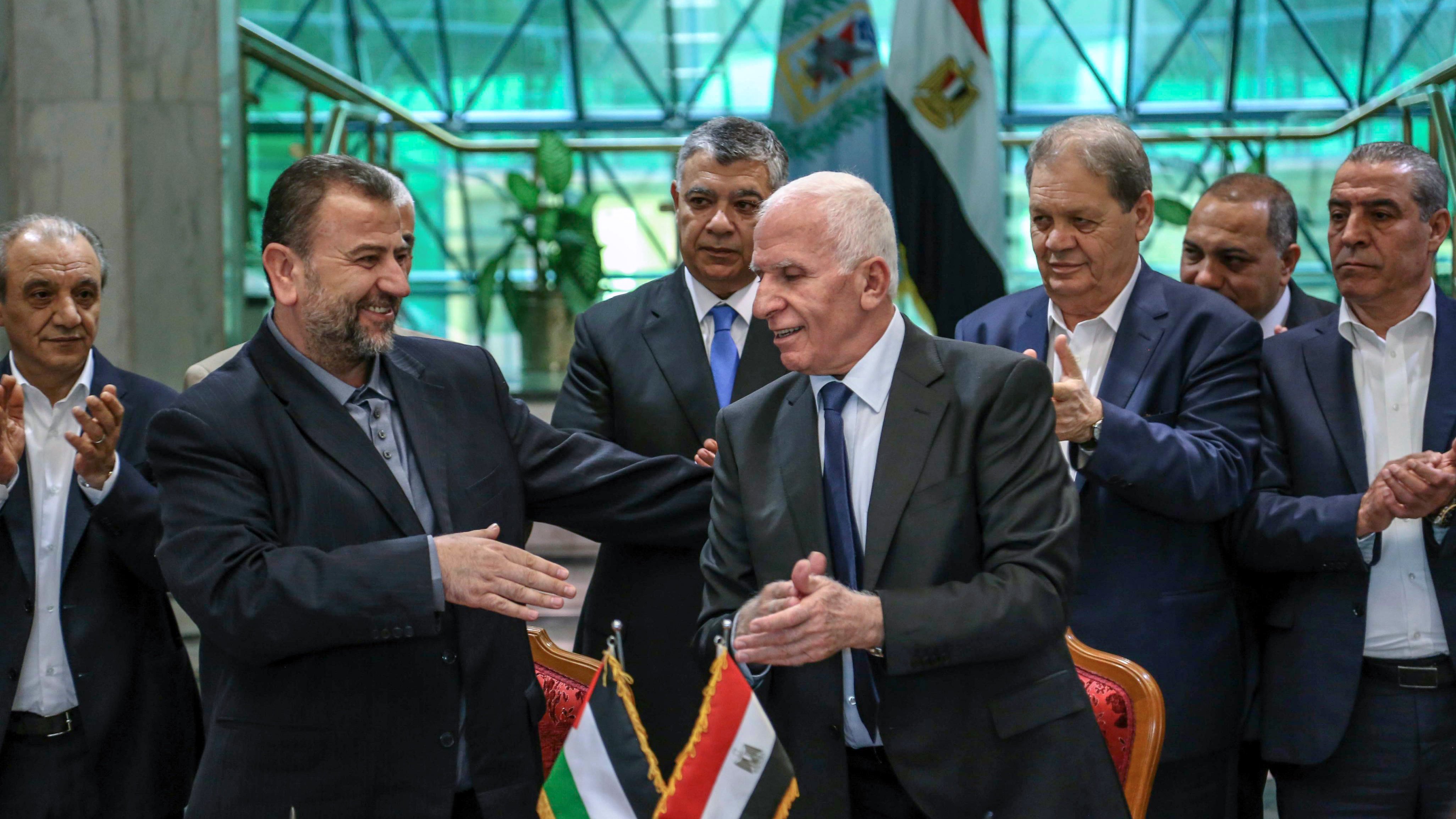 الصورة تظهر مجموعة من الشخصيات رجالية، تتواجد في مكان رسمي، حيث يتم عقد اتفاق أو توقيع. في المقدمة، يظهر شخصان shaking hands، يعبر ذلك عن اتفاق أو تعاون. الخلفية تحتوي على علمين: علم فلسطين وعلم مصر، مما يدل على موضوع النقاش أو الحدث. باقي الأشخاص في الصورة يبدون ممتنين أو يدعمون اللحظة، مما يعكس أهمية الحدث.