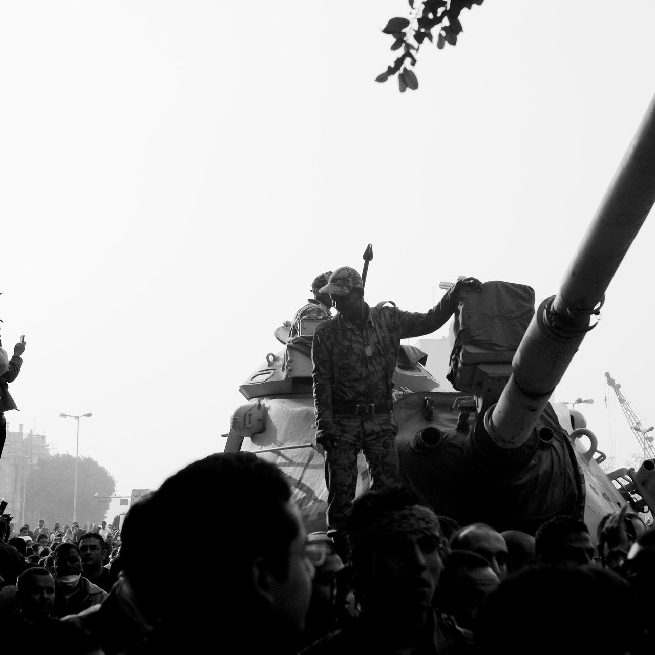 تظهر الصورة مشهداً من مظاهرة أو حدث احتجاجي، حيث يوجد عدد كبير من الناس يتجمعون حول دبابة. أحد الجنود يقف فوق الدبابة ويرفع يدَه كما لو كان يحمل شيئاً أو يجذب الانتباه. الأجواء تشير إلى توتر أو حالة من الحماس بين الحشود. الصورة تُظهر تبايناً كبيراً بين الأضواء والظلال، مما يضفي عليها طابعاً دراماتيكياً.
