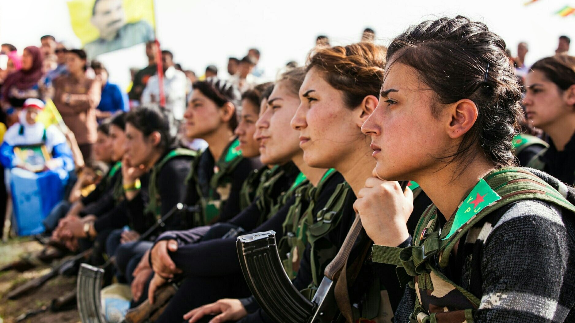 L'image montre un groupe de femmes assises en rang, portant des uniformes militaires avec des armes à leurs côtés. Elles semblent attentives et réfléchies, regardant vers l'avant. En arrière-plan, on peut apercevoir d'autres personnes réunies, ainsi qu'un drapeau symbolique. L'ambiance semble être celle d'un rassemblement ou d'une cérémonie.