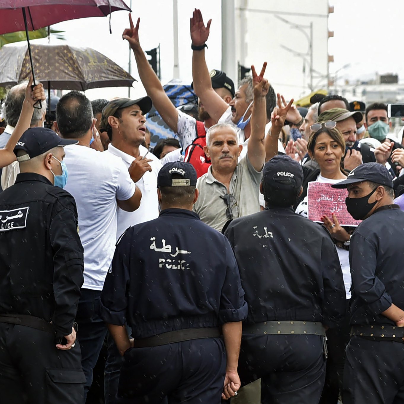 L'image montre une scène de tension entre un groupe de manifestants et des policiers. Les manifestants, certains avec des parapluies, lèvent les bras et expriment leurs opinions. Les policiers, en uniforme et portant des masques, se tiennent en face d'eux, créant un contraste clair entre les deux groupes. La situation semble chargée d'émotion, avec des personnes qui cherchent à faire entendre leur voix. Le temps est pluvieux, ajoutant une atmosphère dramatique à l'événement.