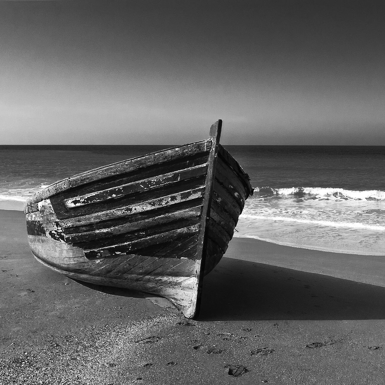 L'image montre un bateau en bois qui est échoué sur une plage. Le bateau, usé par le temps, est orienté vers la mer. La plage est accompagnée de vagues qui se brisent doucement sur le rivage. Le contraste en noir et blanc donne une atmosphère calme et mélancolique à la scène, mettant en valeur les textures du bois et les formes des vagues. Le ciel est dégagé, ajoutant à la sérénité de l'image.