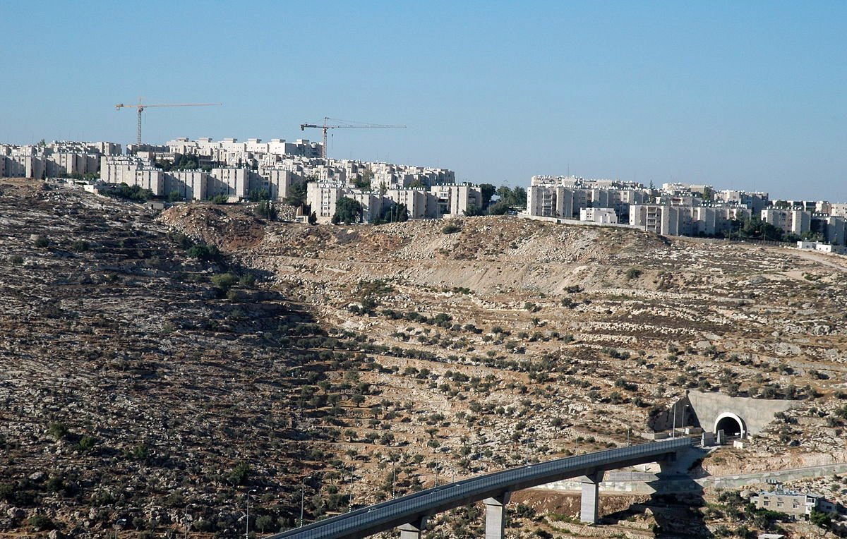 L'image montre un paysage caractérisé par une colline aride et rocheuse, sur laquelle se trouvent des bâtiments modernes. En arrière-plan, on peut voir des constructions en cours, avec des grues qui ajoutent à l'atmosphère de développement urbain. Au premier plan, une structure en béton est visible, semblant faire partie d'une voie ou d'un pont qui traverse le terrain. Le ciel est dégagé, ce qui donne une impression de luminosité et d'espace.