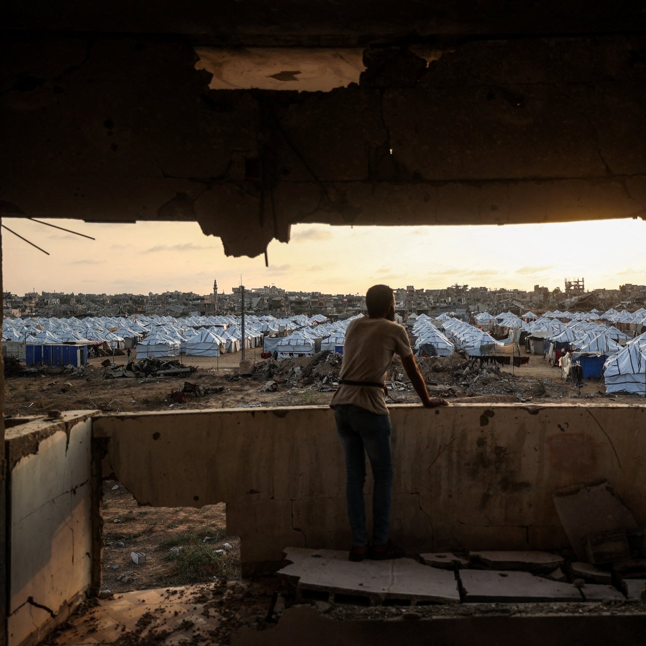پسری از لبه‌ی یک ساختمان ویران به سمت اردوگاه چادرها در غروب نگاه می‌کند.
