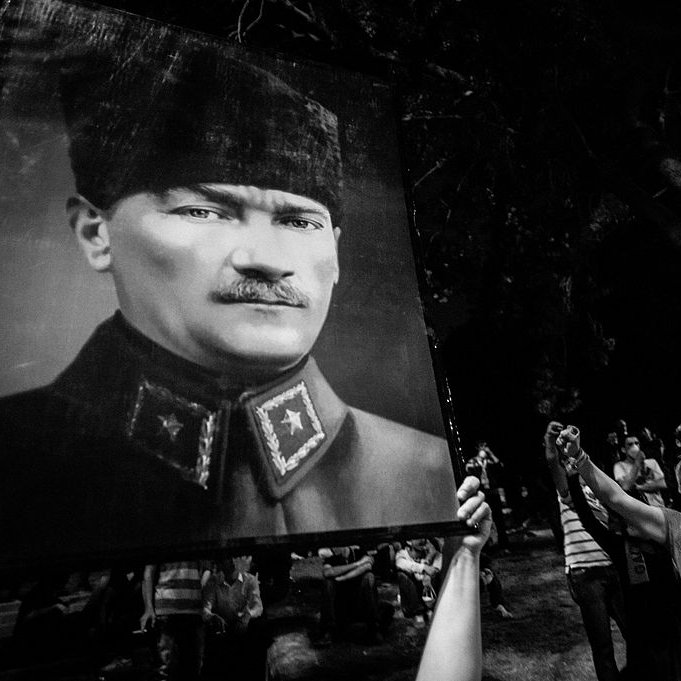 L'image montre un rassemblement nocturne où des gens tiennent un grand portrait d'une figure historique. Les participants semblent exprimer leur admiration ou leur respect, levant le poing en signe de solidarité ou de protestation. L'atmosphère est festive mais aussi solennelle, avec des ombres et une luminosité faible qui accentue l'intensité du moment.