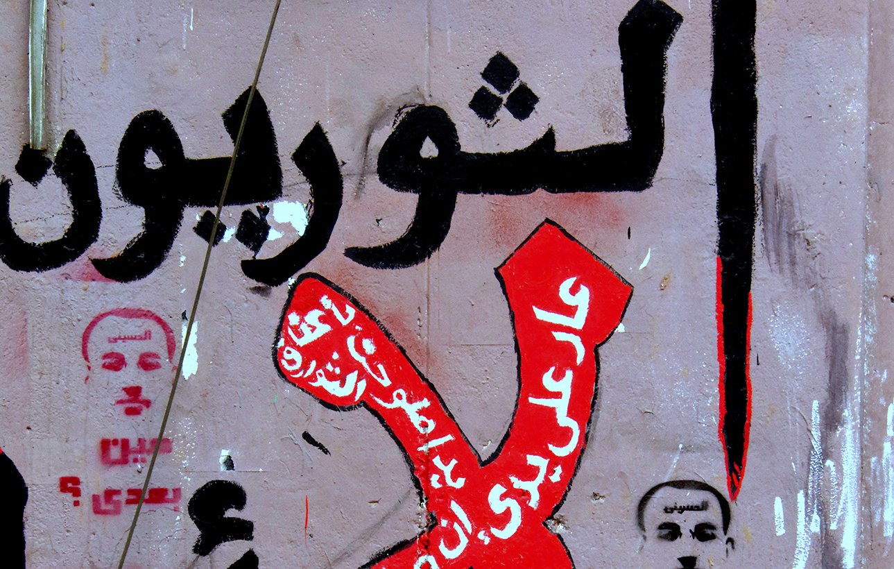 L'image montre un mur recouvert de graffitis, avec des inscriptions en arabe. On peut y voir le mot "Non" stylisé en rouge, entouré de phrases opposées à certaines idées ou actions. Des visages ou portraits, probablement des figures publiques, sont également esquissés. L'ensemble de la composition véhicule un message fort et engagé, typique de la communication visuelle dans des contextes de protestation sociale ou politique.