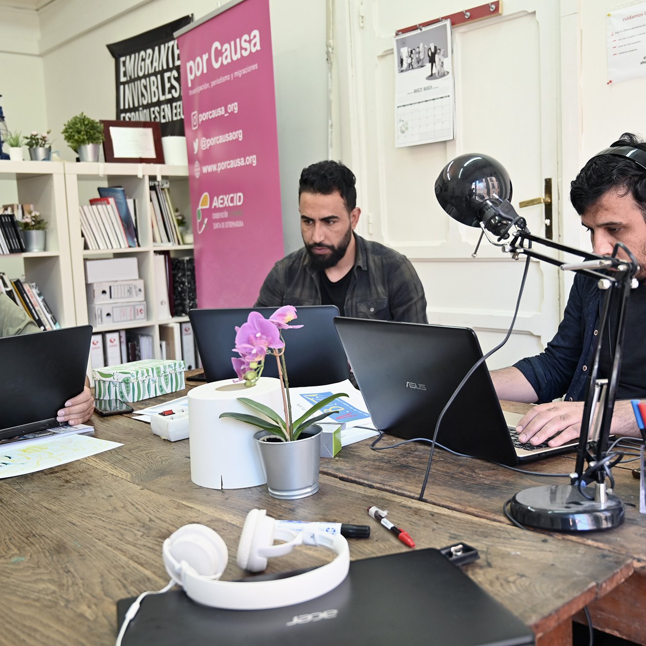 L'image montre trois hommes assis autour d'une table en bois, chacun travaillant sur un ordinateur portable. Ils semblent concentrés sur leurs tâches. Sur la table, on peut voir des objets tels qu'un casque, des stylos et une plante en pot. À l'arrière-plan, il y a des affiches et des livres, ce qui donne une ambiance de bureau ou d'espace de travail collaboratif. Un microphone est également visible sur la table, ce qui pourrait indiquer qu'ils sont en train d'enregistrer ou de diffuser quelque chose.