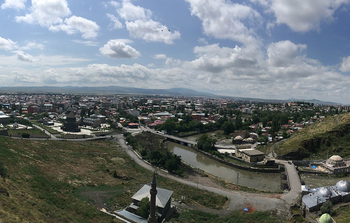 L'image représente une vue panoramique d'une ville avec un paysage verdoyant. On peut voir des toits de maisons, des bâtiments et des infrastructures qui s'étendent à travers la vallée. Le ciel est partiellement nuageux, laissant passer la lumière du soleil. Au premier plan, il y a des collines et des routes qui serpentent à travers le paysage. L'ensemble dégage une atmosphère paisible et naturelle, avec une harmonie entre l'urbanité et la nature environnante.
