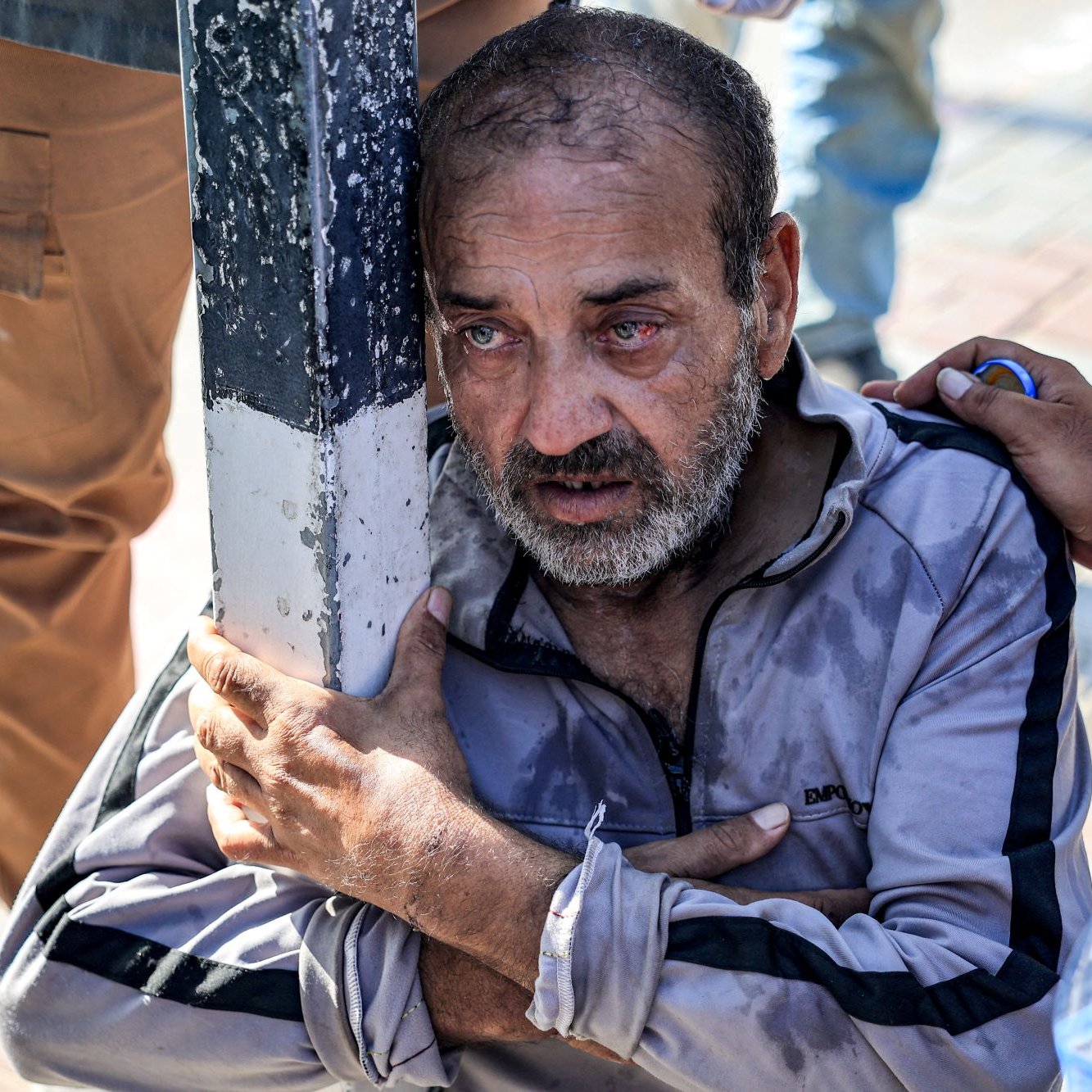 L'image montre un homme assis près d'un poteau, avec une expression de détresse sur son visage. Il semble bouleversé, les yeux plissés comme s'il était en larmes. Il porte une tenue décontractée, légèrement sale et mouillée. Ses bras sont enroulés autour de lui, exprimant une posture de vulnérabilité et de désespoir. À proximité, on peut voir une bouteille d'eau, suggérant qu'il pourrait être en situation difficile ou de précarité. L'environnement autour semble urbain, avec des pavés au sol.