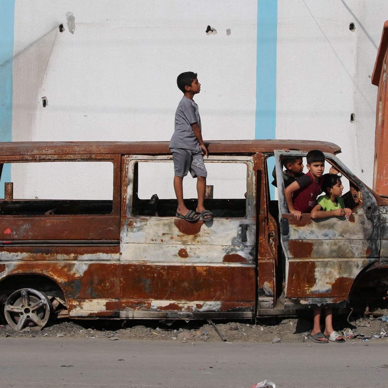 L'image montre un groupe d'enfants jouant autour d'une vieille fourgonnette rouillée et endommagée. Un enfant est debout sur le toit de la voiture, tandis que d'autres sont assis et debout autour d'elle, semblant s'amuser malgré l'environnement délabré. En arrière-plan, on aperçoit un bâtiment aux murs abîmés. L'atmosphère semble à la fois joyeuse et nostalgique, illustrant la résilience des enfants dans un cadre difficile.