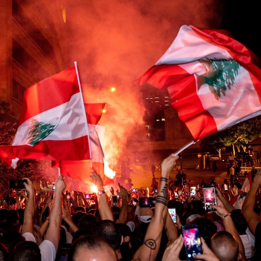 The image depicts a large gathering of people celebrating, likely in an urban setting. Many individuals are holding flags of Lebanon, which feature a red and white design with a green cedar tree. The atmosphere appears vibrant and energetic, with colorful smoke and lights adding to the scene. People are also raising their phones to capture the moment, suggesting a significant event or celebration taking place.